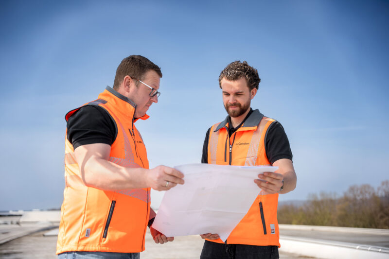 Marcel Möller und Darian Consten stehen auf einem Flachdach und schauen auf einen Plan